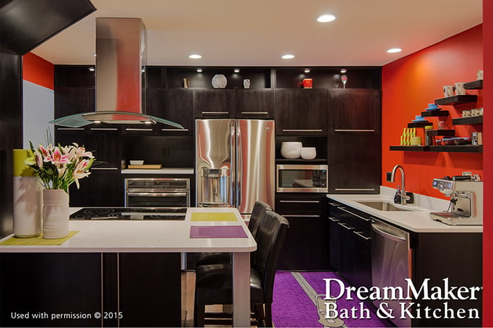 A contemporary-style kitchen, with orange walls, stainless steel appliances, black cabinetry and a purple rug. The DreamMaker® Bath & Kitchen logo is in the lower-right corner.