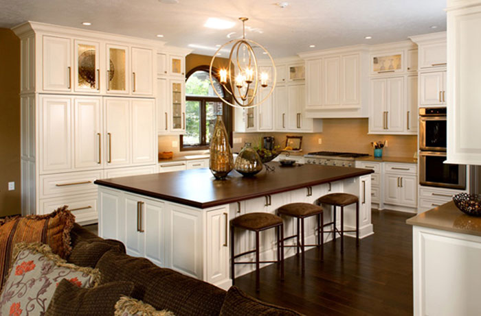 A kitchen with white cabinets including an island with a brown wooden table top. An chandelier hangs above the kitchen island, and a living room couch is partially visible in the foreground.