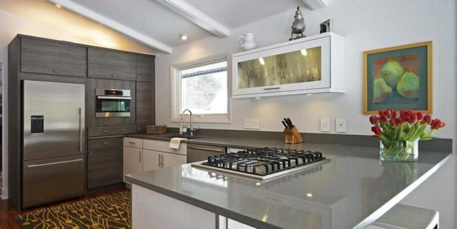 A kitchen remodel showcasing sleek gray countertops, stainless steel appliances and a mix of white and gray wooden cabinets. A painting of pears and a vase full of red flowers are visible at the right side of the frame.