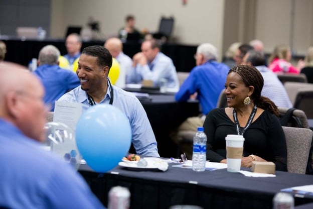 Two smiling franchisees sit at a table in a large conference room. Other attendees are visible in the foreground and background.