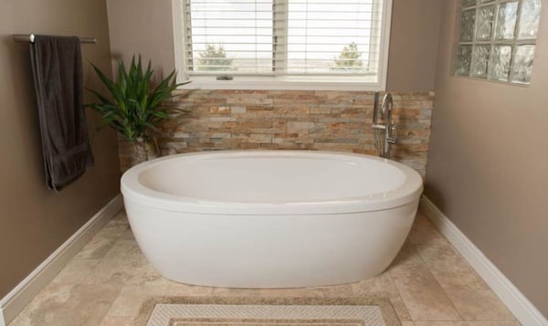 A white ceramic bathtub on beige floor tiles with light brown paint and small, patterned tiles on the back wall beneath a window. A towel and green plant can be seen to the left of the tub.