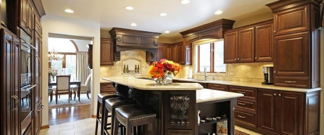 A view of a newly remodeled kitchen, with dark wooden cabinets, a marble backsplash and counters, leather-upholstered stools and a centerpiece of flowers on the kitchen island.