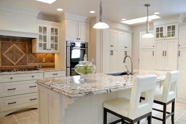 A modern kitchen features white cabinetry, a pale granite-topped center island with two white chairs, and beige large-tile floors. A decorative jar of green apples sits on the island, which also has a sink.