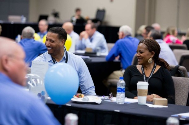 A man and a woman wearing lanyards sitting at a cloth-covered conference table smile at someone off-camera to the left. In the background are several groups of business people at other conference tables.